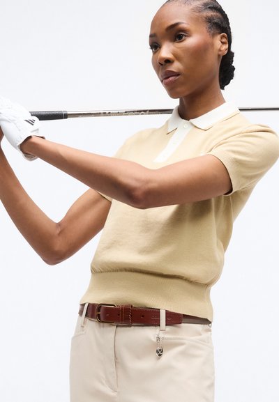 Γυναίκα με μπεζ πόλο και παντελόνι κρατά ρόπαλο γκολφ πίσω από τους ώμους της, φορώντας λευκό γάντι στο αριστερό της χέρι, με φόντο λευκό.