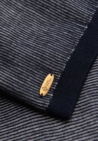 Gros plan sur un tissu tricoté rayé bleu marine et gris avec une petite étiquette métallique ovale portant l'inscription "OMBRE HERITAGE" attachée près d'un bord uni bleu marine.