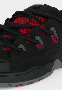 Gros plan d’une chaussure de sport noire et rouge avec des lacets noirs, un tissu en mesh rouge et une semelle en caoutchouc grise et rouge.