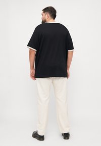 Zwarte katoenen t-shirt met witte mouwdetails, gecombineerd met crèmekleurige broek, gezien vanuit de achterkant. Model staat tegen een eenvoudige witte achtergrond.