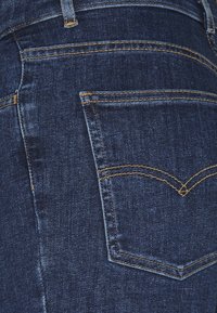 Primer plano de un bolsillo trasero de unos pantalones vaqueros de mezclilla azul oscuro con costuras amarillas y detalles de la trabilla del cinturón.