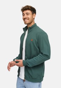 Sweat-shirt zippé vert avec texture côtelée, col montant et un petit logo en cuir, assorti à un t-shirt blanc et un jean clair.