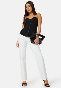 Top nero senza spalline con bottoni e tasche, abbinato a jeans bianchi a gamba dritta. La modella tiene una borsa nera a trama e indossa tacchi neri.