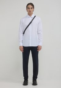 Camicia bianca con colletto a punta, abbinata a pantaloni blu navy. Il modello indossa scarpe nere e una borsa a tracolla nera.
