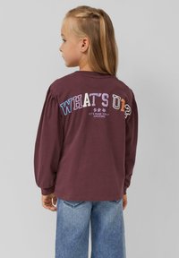 Weinrotes Langarmshirt mit Puffärmeln, das auf der Rückseite mehrfarbigen Text trägt: „WAS GIBT'S?“ und „LASS UNS DEN TAG EINZIGARTIG MACHEN.“