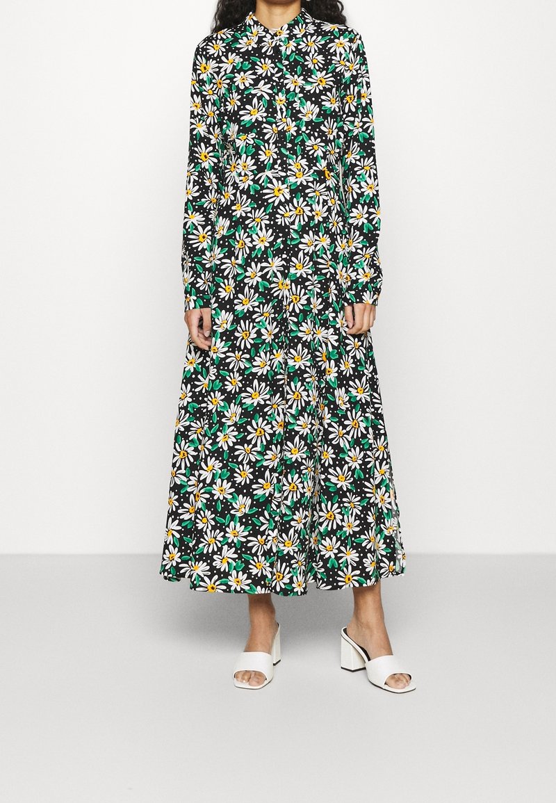 Femme portant une longue robe noire avec des imprimés de marguerites blanches et des feuilles vertes, associée à des sandales blanches à talons blocs et bouts ouverts.