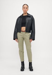 Jaqueta curta de couro sintético preto com mangas largas e fecho frontal, combinada com calças verde-oliva e botas de tornozelo pretas.