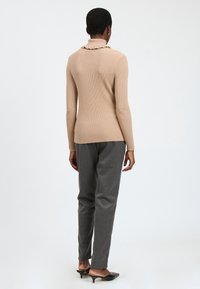 Persona in piedi di spalle che indossa un maglione beige a costine con collo alto, pantaloni grigi su misura e mules nere con tacco su uno sfondo bianco.