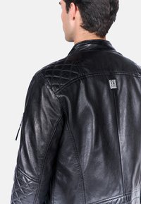 Czarna skórzana kurtka z pikowanym wzorem na ramionach, gładką teksturą i kieszenią na zamek na rękawie, z metką marki z tyłu.