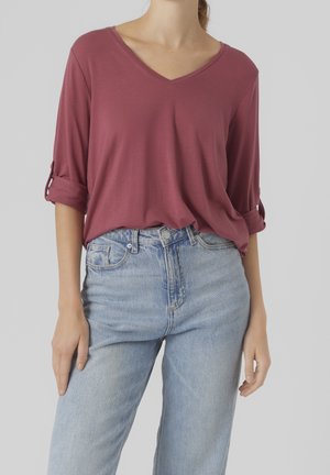  Mujer con una camiseta de manga remangada, de color malva y escote en V, metida dentro de unos vaqueros de talle alto y azul claro, sobre un fondo liso.