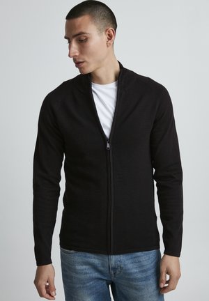 Svart glidelåsgenser med ribbet tekstur og høy krage, sammen med en hvit T-skjorte og blå jeans. Tettsittende silhuett.