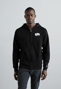 Sort lynlås hoodie med hætte, med et hvidt logo på venstre bryst. Lavet af blødt, tekstureret stof, som er parret med mørkt stribede bukser.