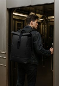 Mann mit schwarzem Rolltop-Rucksack, der in eine U-Bahn einsteigt, die Türgriff hält und eine dunkle Jeansjacke sowie Hose trägt.