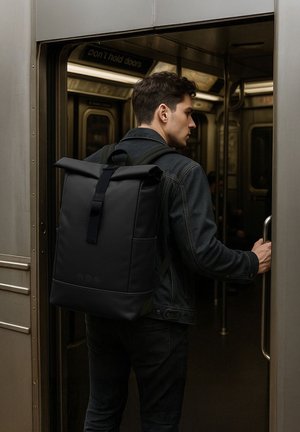 Homme avec un sac à dos noir à rabat entrant dans un train de métro, tenant la poignée de la porte, portant une veste en denim foncé et un pantalon.