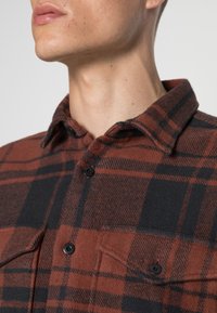 Flanelová košile v barvě spálené oranžové a černém kostkovaném vzoru. Má černé knoflíky a náprsní kapsu. Vyrobena z měkké, texturované tkaniny.
