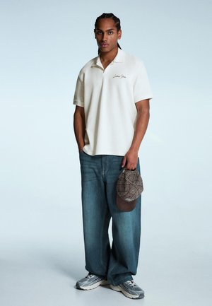 Bílé polo tričko s krátkým rukávem a vyšitým logem, doplněné volnými modrými džíny. Drží hnědou kostkovanou čepici, na sobě má šedé tenisky.