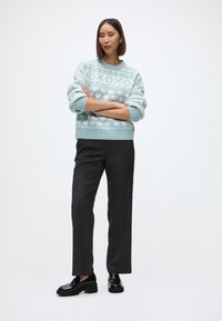 Gaiši zils džemperis ar sirdīšu rakstu, apvienots ar tumšiem, piegrieztiem biksēm. Modelis valkā melnas apavus ar masīvu zoli. Tīra, minimāla fona.