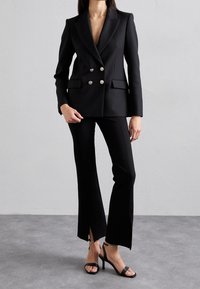 Blazer doppiopetto nero su misura con bottoni dorati, abbinato a pantaloni a zampa coordinati con spacchi e sandali con tacco alto neri.