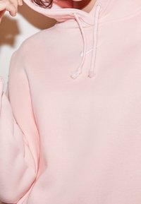 Sudadera rosa claro hecha de tela suave, con capucha ajustable y un pequeño logo blanco. El diseño incluye detalles de puños y dobladillo acanalados.
