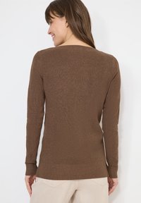 Femme portant un pull en tricot à manches longues de couleur marron, souriante, de dos, vêtue d'un pantalon beige clair sur un fond blanc uni.