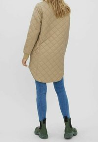 Manteau long matelassé beige avec un ourlet arrondi, associé à un jean bleu ajusté et des bottines vertes. Le manteau comporte un col montant et des poignets côtelés.