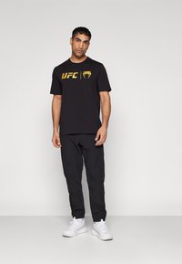 Sort bomulds T-shirt med et gult UFC-logo, parret med sorte bukser og hvide sneakers. Enkel design med korte ærmer.