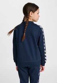 Felpa blu navy con colletto alto, caratterizzata da strisce diagonali bianche lungo le maniche. Il tessuto sembra morbido e testurizzato.