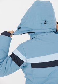 Blå isoleret jakke med hætte, med marineblå og lyseblå striber på ærmerne og lynlås ved halsen. Teksturen ser glat ud.