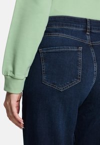 Ciemnoniebieskie dżinsy z pięcioma kieszeniami, średnim stanem, kontrastującym przeszyciem i miękką fakturą denimu. Na górze znajduje się jasnoniebieska bluzka.