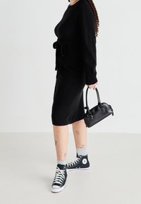 Robe en tricot noire avec une ceinture, portée avec des baskets montantes noires, des chaussettes grises et un petit sac à main noir. Texture lisse, design longueur cheville.