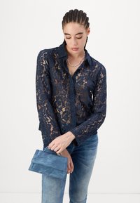 Chemisier en dentelle bleu marine à manches longues, avec un motif floral et une légère transparence. Associé à une pochette en daim bleu.