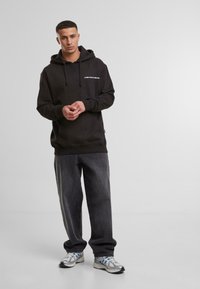 Black hoodie with a front pocket, featuring white text. Paired with dark gray jeans and white/silver athletic shoes. Smooth texture.