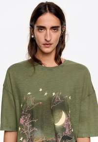 Olijfgroene oversized t-shirt van zacht materiaal met een grafische print van vogels, sterren en een maan in roze en beige tinten.