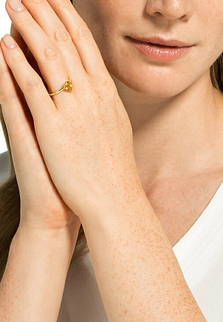Goldring an einer Hand mit leichten Sommersprossen; der Ring hat eine herzförmige Edelsteinfassung, mit einem glatten Band und glänzendem Finish.