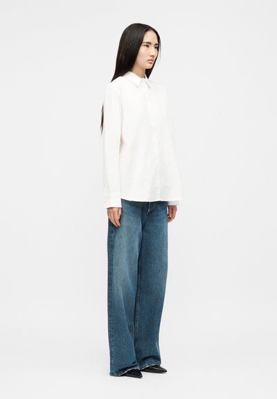 Kvinne som står iført en hvit langermet skjorte med krage, vidbukser i blått denim og svarte spisse sko mot en ensfarget bakgrunn.