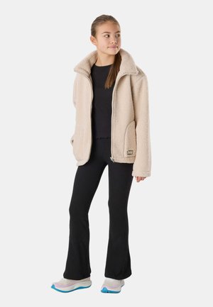 Młoda kobieta stoi, ubrana w beżową polarową bluzę na zamek, czarny prążkowany top i spodnie oraz białe trampki z niebieskimi podeszwami, patrzy w prawo.