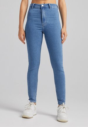 Femme portant un jean skinny taille haute bleu clair et des baskets blanches, debout devant un fond uni gris clair.