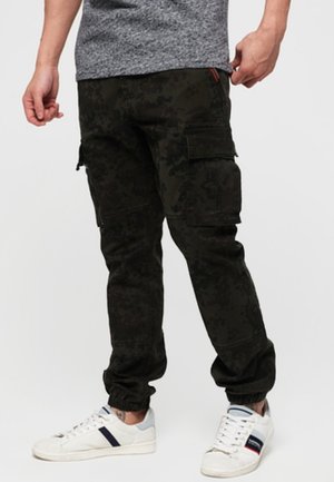 Mężczyzna noszący ciemne spodnie bojówki w kamuflażu z wieloma kieszeniami, w połączeniu z białymi tenisówkami z czarnymi i czerwonymi paskami.