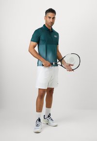 Teal színű póló, amelyen átmenetes csíkok és apró pöttyök vannak, fehér sorttal párosítva. A játékos teniszütőt tart a kezében. A sportcipő kék és sárga részletekkel rendelkezik.