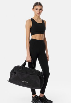 Mujer con ropa deportiva negra sosteniendo una gran bolsa de gimnasio negra con el logo "Cardio Bunny", de pie contra un fondo blanco liso.