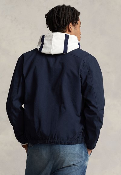 A person with dreadlocks wears a navy jacket with a white hood and navy stripes, facing away against a textured light background.