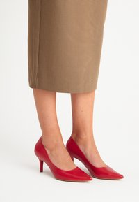 Zapatos de tacón alto de cuero rojo con punta afilada y textura suave, sobre una falda de cuadros marrones.