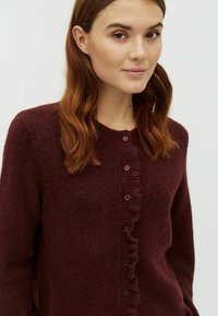 Bordeaux cardigan vyrobený z mäkkého pleteného materiálu má okrúhly výstrih, zapínanie na gombíky a volánový okraj pozdĺž prednej časti.