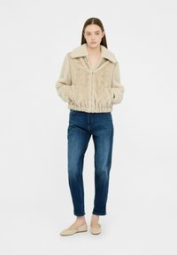 Giacca in pelliccia sintetica beige con chiusura a zip, motivo a treccia testurizzato e ampio colletto. Abbinata a jeans blu e scarpe slip-on chiare.