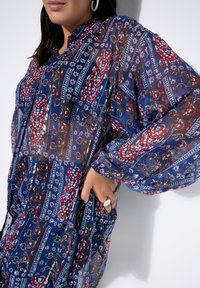 Mujer vestida con una blusa azul transparente con patrones florales en rojo y blanco, mangas largas abullonadas y pendientes de aro plateados, con la mano descansando en la cadera.