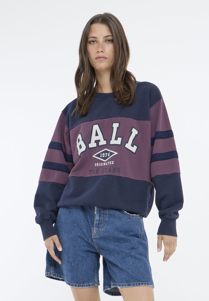 Hanorac colorat în bleu marin și burgundy cu grafică "BALL 1974", cu manșete cu nervuri și o croială lejeră, purtat cu pantaloni scurți din denim albastru.