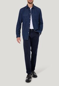 Donkerblauw denim shirt met knoopsluiting aan de voorkant, twee borstzakken en lange mouwen. Gecombineerd met marineblauwe broek en zwarte schoenen.