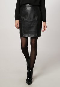 Sort læder mini-nederdel med en glat tekstur, lige skæring og enkelt design. Har en elastisk talje og er stylet med sorte strømpebukser og hæle.