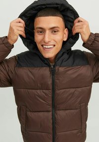 Pruun puff-jakk musta kapuutsiga, millel on tikitud disain, tõmblukk ees ja küljetaskud. Sile tekstuur, kerge materjal.