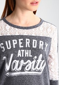 Graues Oberteil mit Spitzenakzenten, das weißen Siebdrucktext mit der Aufschrift "SUPERDRY ATHL VARSITY" in fetter, lässiger Schriftart trägt.
